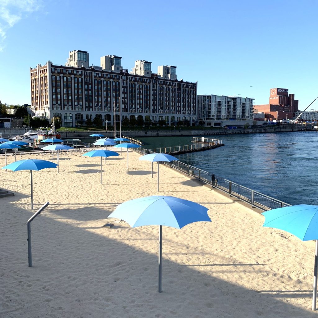 Playa urbana Clock Tower Beach en Montreal con arena y sombrillas azules junto al canal