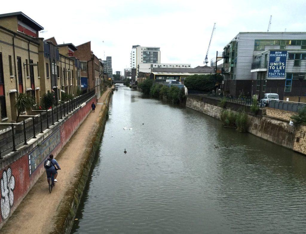 Canal en el este de Londres con edificios industriales, un ciclista y patos nadando en el agua