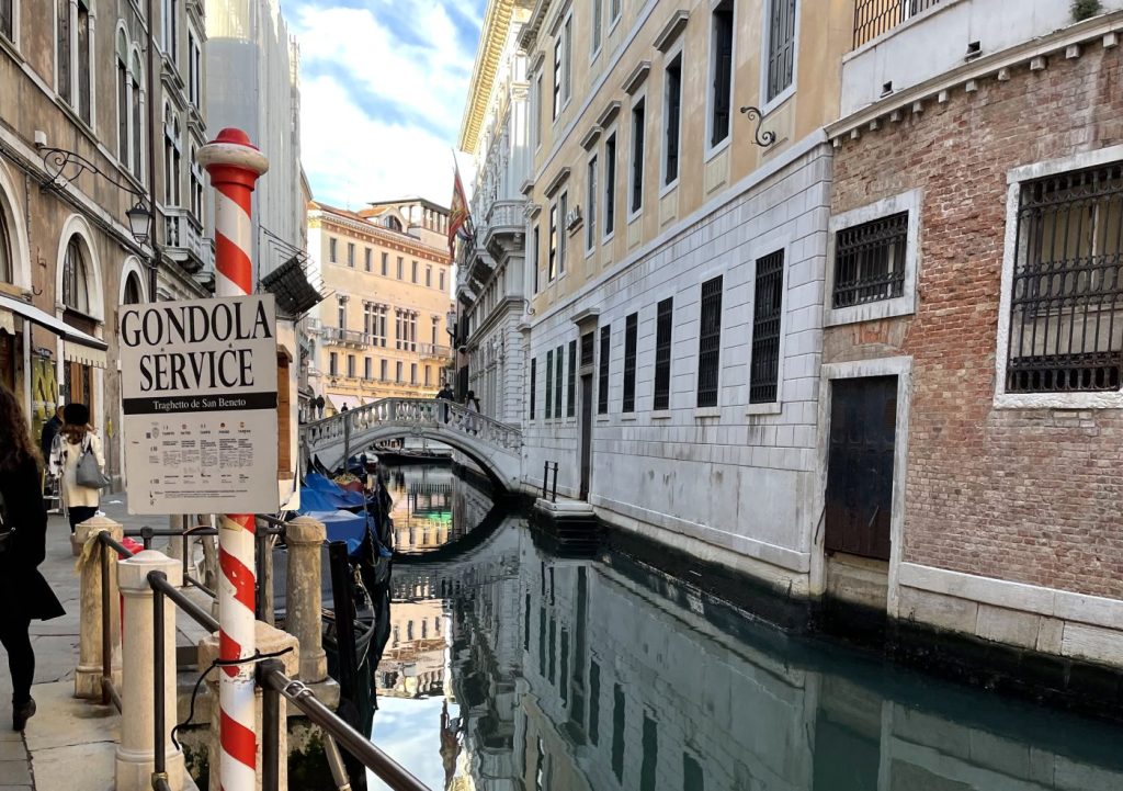 Canal secundario en Venecia con góndolas atracadas y cartel de servicio de góndolas en primer plano